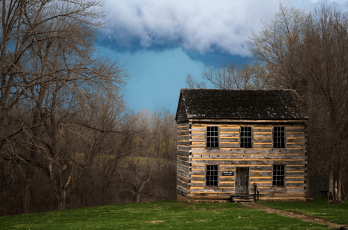 A rustic log cabin stands in a grassy area, with dark storm clouds looming in the background and trees surrounding it.