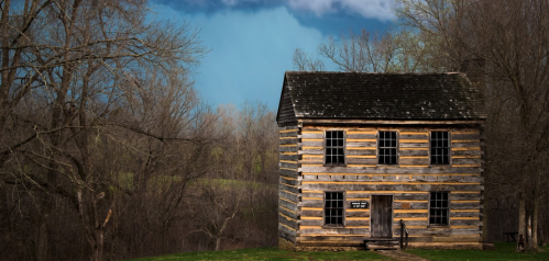 A rustic wooden cabin stands alone against a dramatic sky, surrounded by bare trees and rolling hills.