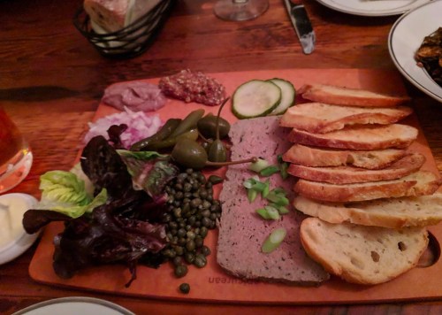 A wooden platter with sliced bread, pâté, pickles, capers, greens, and condiments, set on a rustic table.