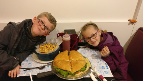 Two kids with glasses smile at the camera, sitting at a table with a large sandwich and a bowl of fries.