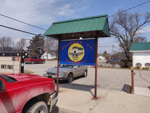Sign reading "Welcome to Murdo's" with a green roof, parked cars, and a small town background.