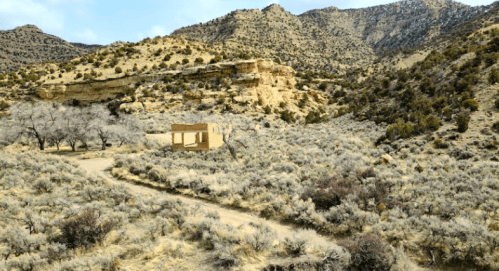 A deserted building surrounded by sparse vegetation and mountains under a cloudy sky.