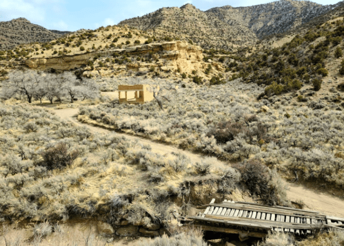 A desolate landscape featuring a dirt road, an old building, and a wooden bridge surrounded by mountains and sparse vegetation.