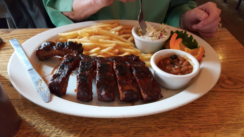 A plate of ribs with fries, coleslaw, baked beans, and an orange slice, with a hand holding a fork.