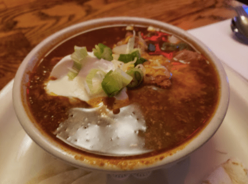 A bowl of chili topped with sour cream, green onions, and cheese, served on a wooden table.