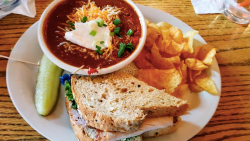 A plate with a sandwich, a bowl of chili topped with cheese and sour cream, potato chips, and a pickle on the side.