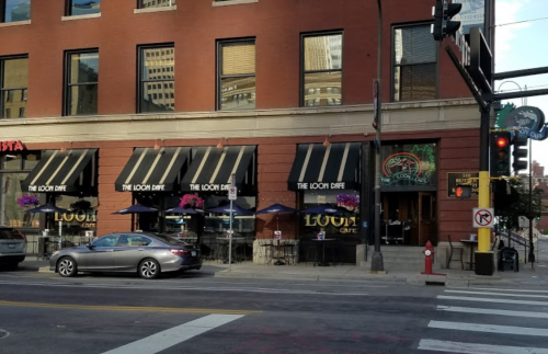 Exterior of The Loom Cafe with outdoor seating, large windows, and flower baskets, located on a city street corner.