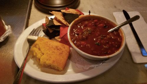 A plate with chili, cornbread, and a mix of red and black tortilla chips, served with a fork and knife.