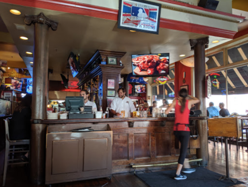 A lively restaurant interior with a bar, staff serving, and a woman walking by, featuring colorful neon signs and TVs.