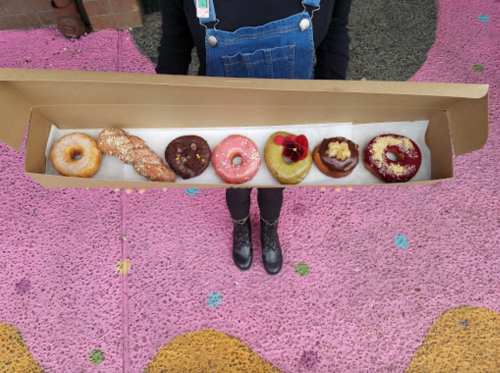 A person holding a box of eight colorful donuts on a pink, textured surface.