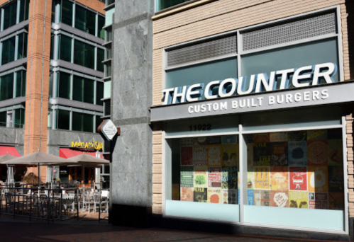 Exterior view of The Counter restaurant with a colorful window display, next to a patio and another eatery.