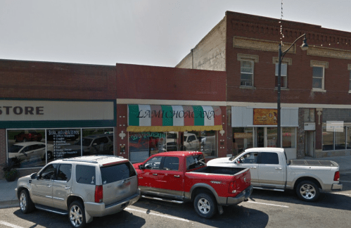 A street view of a small restaurant with a green awning, flanked by parked cars and nearby storefronts.