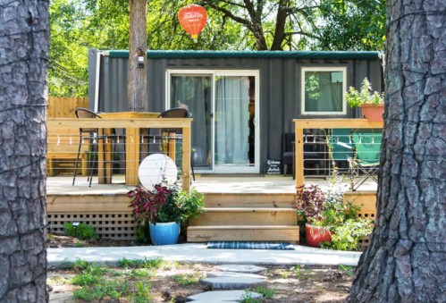 A modern container home with a wooden deck, surrounded by trees and colorful potted plants.
