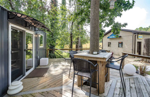 A wooden deck with a table and chairs, surrounded by trees and a building in the background.