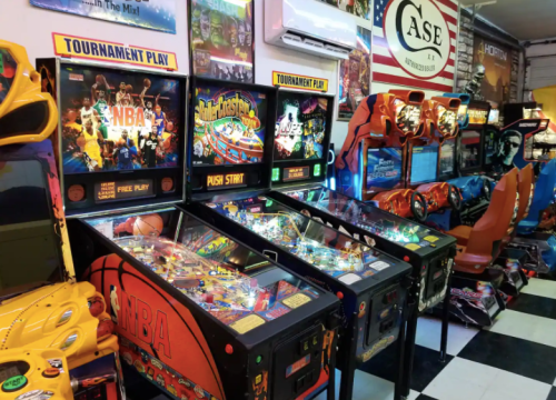 A colorful arcade featuring various pinball machines and video games, with vibrant lights and retro decor.