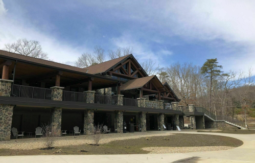 A rustic lodge with stone and wood architecture, surrounded by trees and a clear sky. Outdoor seating is visible.