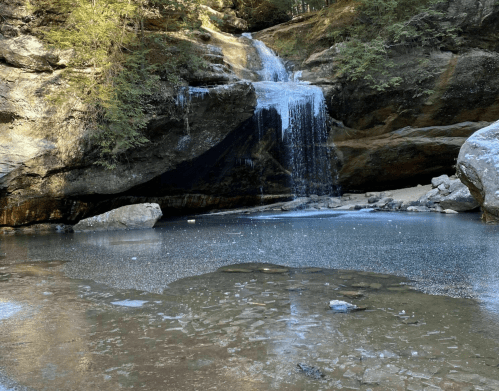 A serene waterfall cascading over rocks into a calm, icy pool surrounded by trees and rugged terrain.