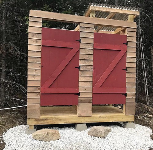 A rustic outdoor structure with red double doors, set on a gravel base surrounded by trees.