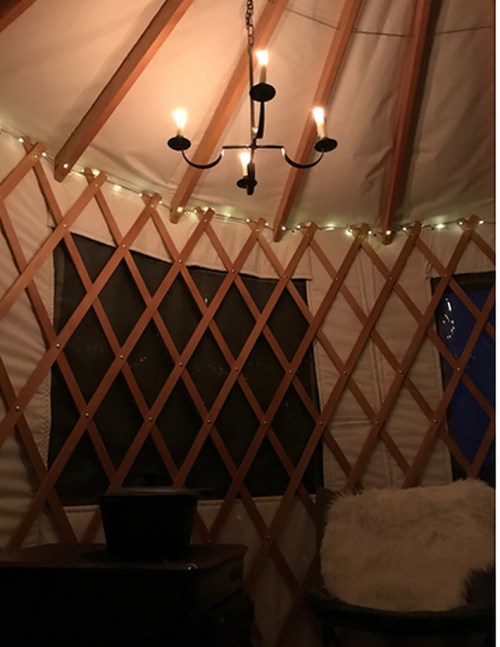Cozy interior of a yurt with a chandelier, soft lighting, and a plush chair against a wooden lattice wall.