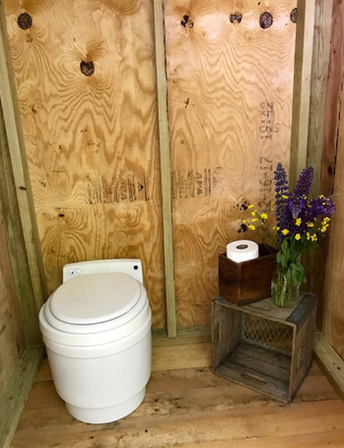 A simple bathroom with a toilet, a wooden crate, a roll of toilet paper, and a vase of flowers on the floor.