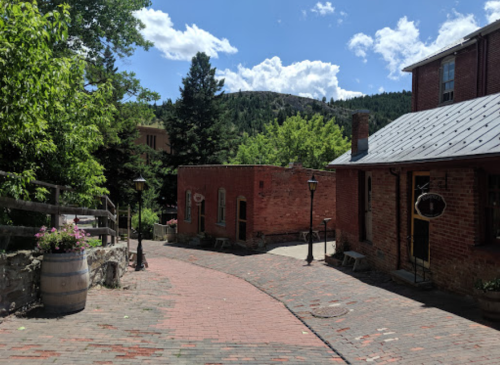 A scenic view of a brick pathway winding through historic buildings, surrounded by trees and mountains under a blue sky.