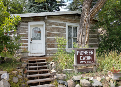 A rustic log cabin with a white door, surrounded by greenery and a sign reading "Pioneer Cabin 1864."