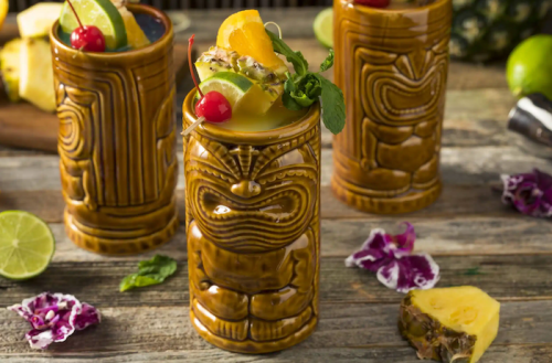 Three tiki mugs filled with colorful tropical cocktails, garnished with fruit and mint, on a rustic wooden surface.