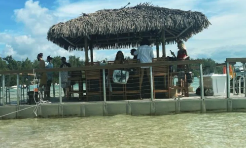 A thatched-roof bar on a pier with people enjoying drinks by the clear water and blue sky.