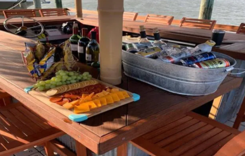 A wooden table with snacks, cheese, grapes, and drinks in a metal tub, set by the water.
