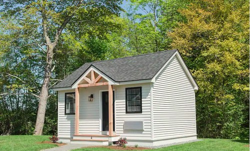A small, modern house with a peaked roof, front porch, and surrounded by lush green trees.