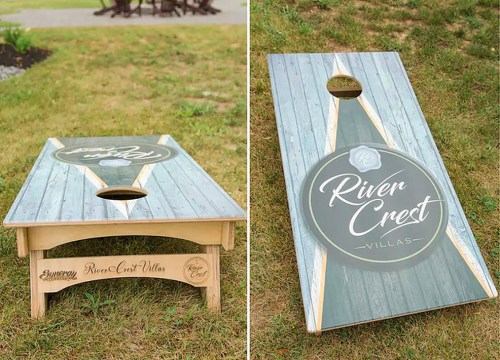 Two cornhole boards on grass, one showing a logo for "River Crest Villas" and the other with a wooden design.