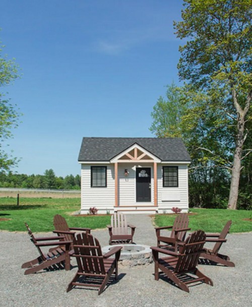 A small house with a porch surrounded by Adirondack chairs and a fire pit, set in a green landscape under a clear blue sky.