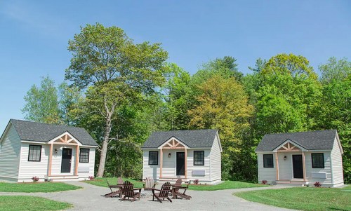 Three cozy cabins surrounded by trees, with a fire pit and seating area in front, under a clear blue sky.