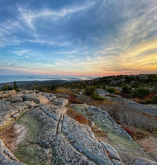A scenic view of rocky terrain with trees, overlooking a coastline at sunset, featuring colorful clouds in the sky.