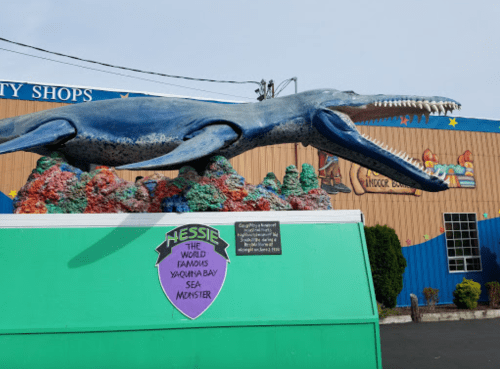 A large blue whale statue with sharp teeth, labeled "Hessie, the world famous Yaquina Bay sea monster," in front of a colorful building.