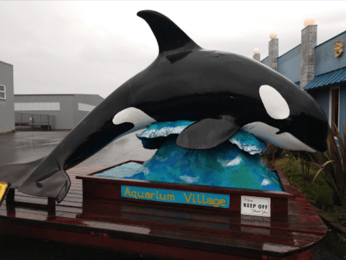 A large orca sculpture on a colorful base outside Aquarium Village, with a "Keep Off" sign nearby.