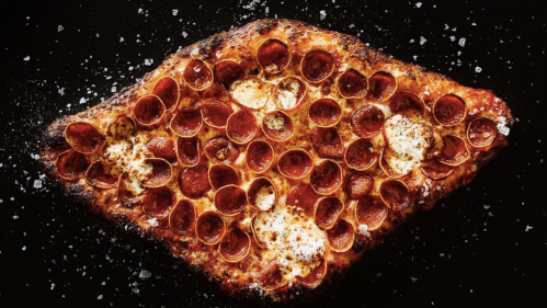 A close-up of a crispy pizza topped with pepperoni and melted cheese, sprinkled with coarse salt on a dark background.