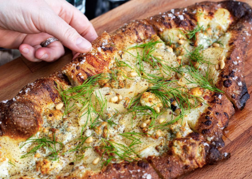 A hand reaches for a slice of pizza topped with cheese, herbs, and pine nuts on a wooden board.