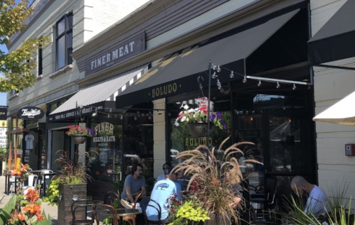A sunny street view of a restaurant with outdoor seating, featuring awnings and colorful flowers.