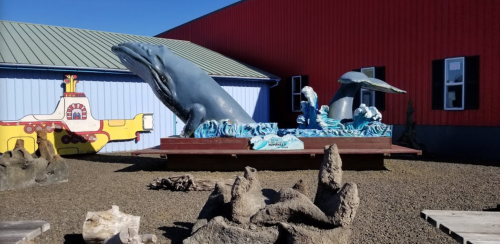 A large whale sculpture with waves, surrounded by colorful buildings and driftwood in an outdoor setting.