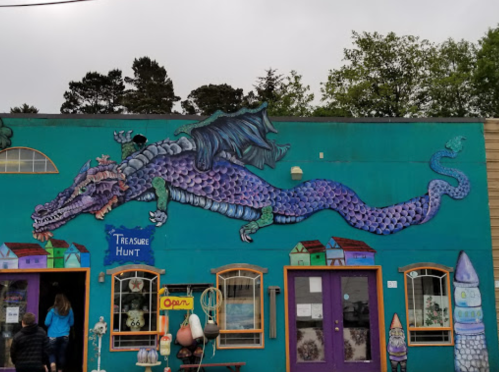 Colorful mural of a dragon on a teal building, with a sign reading "Treasure Hunt" and people walking by.