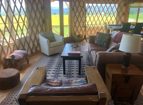 Cozy yurt interior with a leather sofa, armchair, coffee table, and large windows showcasing a scenic view.
