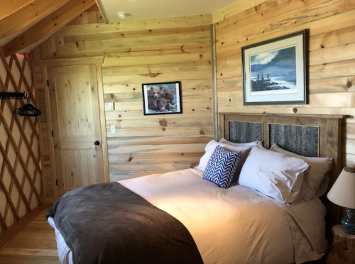 Cozy wooden bedroom with a double bed, decorative pillows, and rustic walls, featuring a framed landscape picture.