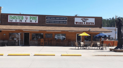 Exterior of Twisted Pine Winery featuring a sign for wine tasting, cheese, and gifts, with outdoor seating and umbrellas.