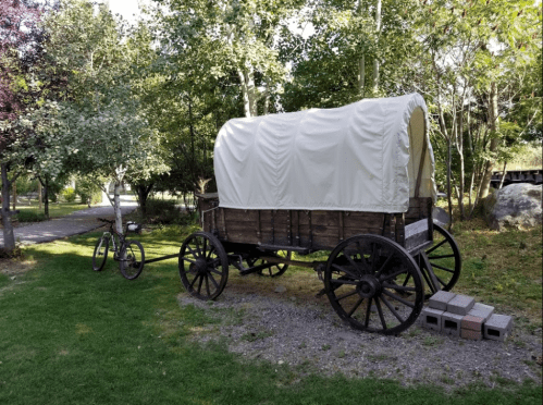 A wooden covered wagon parked beside a bicycle in a green park with trees and a pathway.