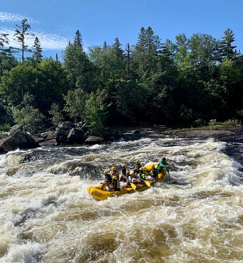 A group of people in a yellow raft navigating through turbulent whitewater rapids surrounded by trees and blue sky.