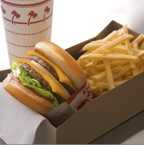 A cheeseburger with lettuce and tomato beside a box of fries and a drink in a paper cup.