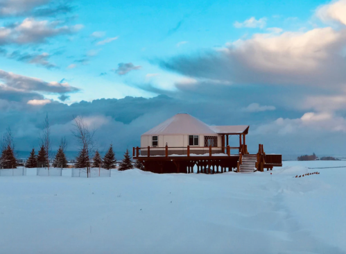 A cozy white house on stilts surrounded by snow, with a blue sky and clouds in the background.