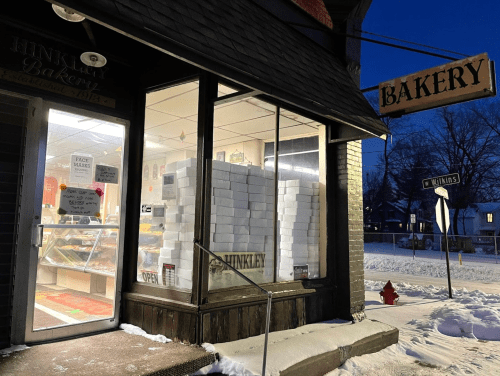 A cozy bakery storefront at dusk, with snow on the ground and a sign reading "BAKERY" hanging above the entrance.