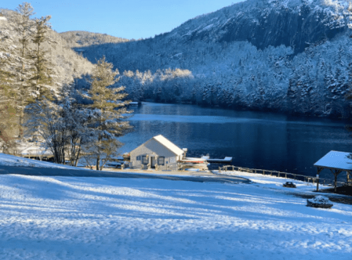 A serene winter scene with a snowy landscape, a calm lake, and a small white cabin surrounded by trees and mountains.